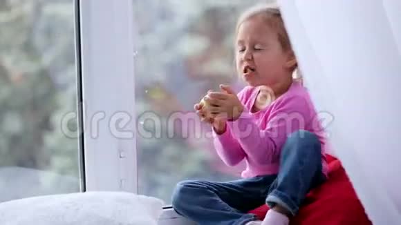 滑稽可爱的小女孩坐在窗台上吃苹果的肖像视频的预览图