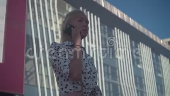 商务女性穿着一件白色衬衫上面挂着豆站在现代办公楼附近打电话视频的预览图