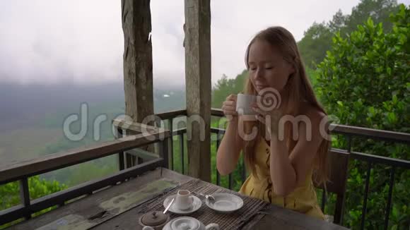一名年轻女子喝茶或咖啡坐在山上的一家咖啡馆里欣赏山景的慢镜头视频的预览图