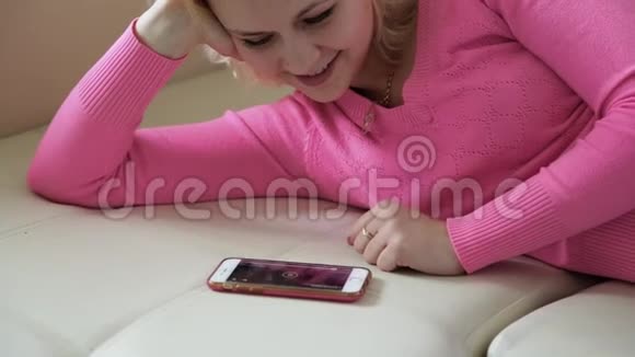 可爱的女人在沙发上放松看着电话微笑着女孩通过互联网交流视频的预览图