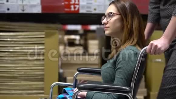 一个年轻的女人坐在轮椅上女助理驾驶一个坐轮椅的女人沿着商店散步视频的预览图