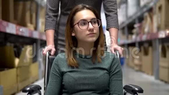 一个年轻的女人坐在轮椅上女助理驾驶一个坐轮椅的女人沿着商店散步视频的预览图