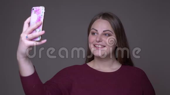 年轻超重的白种人女性在电话中自拍的特写背景是灰色的视频的预览图