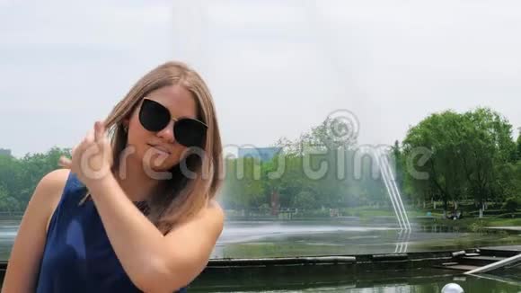 在喷泉附近的公园里一位戴太阳镜时尚发型的美丽年轻女孩的特写肖像可爱视频的预览图