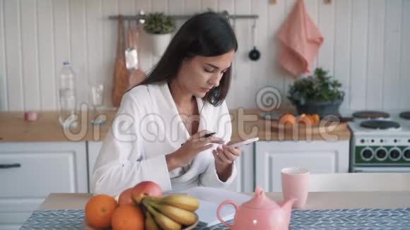 女人用电话坐在厨房在笔记本上写清单慢动作视频的预览图
