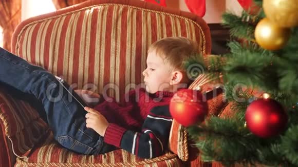 圣诞早晨小男孩躺在扶手椅上在数码平板电脑上玩游戏视频的预览图