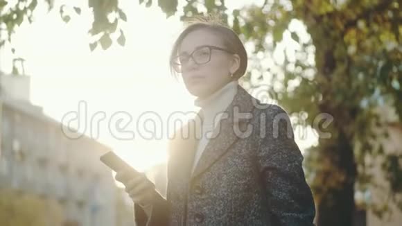 女人在户外使用智能手机戴眼镜和外套的女孩的肖像视频的预览图