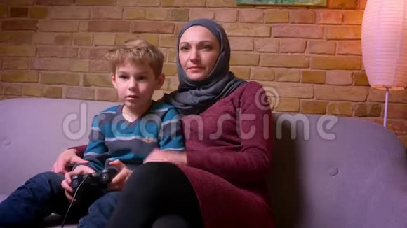 专注的小男孩在玩视频游戏他的穆斯林母亲戴着头巾拿着操纵杆试图在家里帮助他视频的预览图