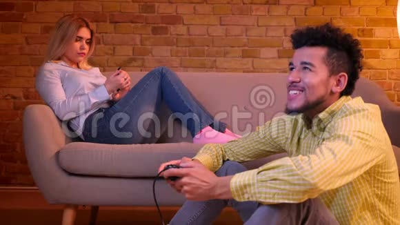 非洲男人坐在地板上快乐地玩电子游戏白种人女孩在舒适的家里等待无聊视频的预览图