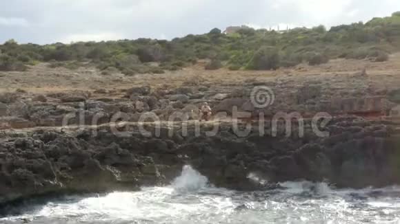 鸟瞰海浪撞击岩石悬崖海岸在阳光明媚的一天与人坐在岩石上马洛卡岛阳光明媚视频的预览图
