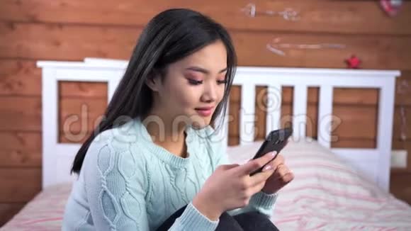 中等特写微笑的年轻亚洲女孩在卧室里坐在床上用智能手机聊天视频的预览图