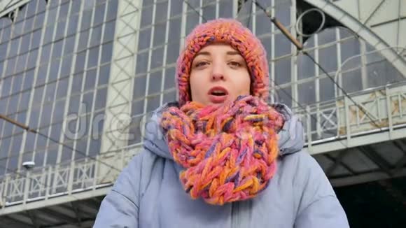 一个年轻漂亮的成年女孩穿着蓝色外套戴着五颜六色的帽子和围巾在阳光明媚的火车站的冬天肖像视频的预览图
