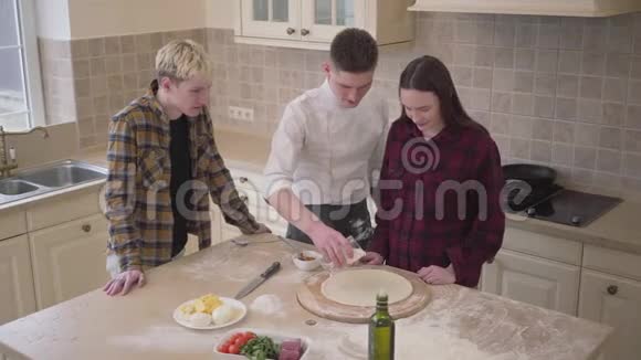 年轻的披萨制造商教他的朋友如何在家里的厨房里做披萨自信的皮萨约罗涂抹奶油和视频的预览图