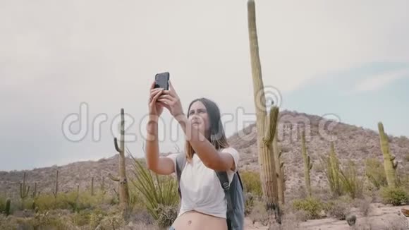 慢动作年轻快乐的女游客拍智能手机照片在史诗国家公园的仙人掌沙漠走向汽车视频的预览图
