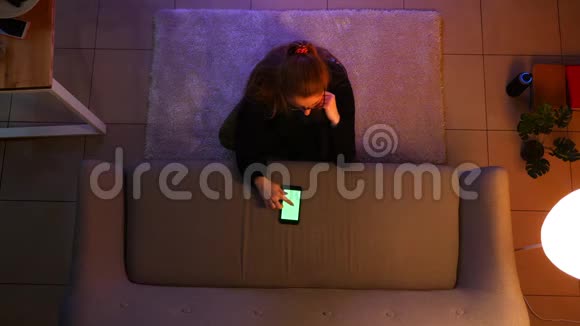 一位漂亮的少女坐在地毯上用绿色屏幕的手机看电视的特写镜头视频的预览图