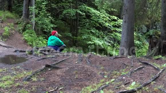 在春天的森林里徒步旅行时一个戴着红帽子和绿松石夹克的年轻人休息的后景库存数据视频的预览图