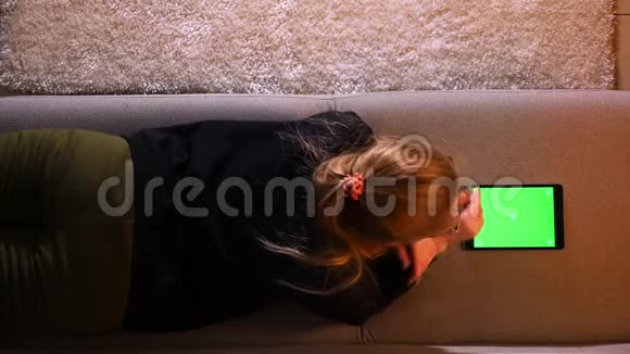 在舒适的室内沙发上躺着的漂亮女孩用绿色屏幕的平板电脑进行近距离的上背部拍摄视频的预览图