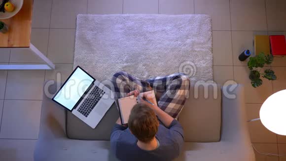 一位年轻的商人在室内坐在沙发上用笔记本电脑做工作和笔记的特写镜头视频的预览图