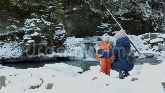 妈妈和儿子坐在雪覆盖的瀑布小山和松林的背景上冬季阴天视频的预览图