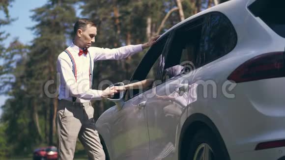 一个穿着白色衬衫吊带和红色领结的时髦小伙子站在车旁亲吻女孩的手视频的预览图