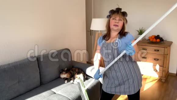 中年妇女穿着围裙和卷发的滑稽家庭主妇用拖把唱歌跳舞用刷子刷在棍子上打扫房子视频的预览图