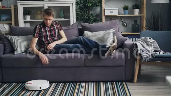 无忧无虑的年轻人打开了机器人吸尘器然后坐在沙发上使用智能手机享受方便视频的预览图