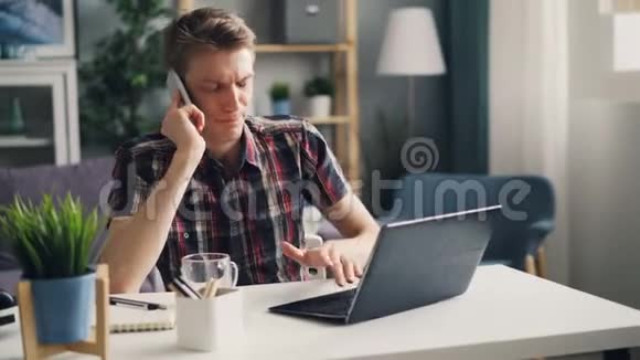 愤怒的年轻人用手机说话用笔记本电脑表达愤怒和愤怒独自在现代公寓工作视频的预览图