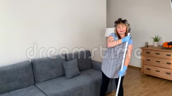 中年妇女穿着围裙和卷发的滑稽家庭主妇用拖把唱歌跳舞用刷子刷在棍子上打扫房子视频的预览图