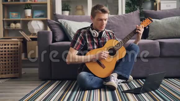 穿休闲服装的年轻人正在看使用笔记本电脑的教程并在家中坐在地板上弹吉他学习视频的预览图