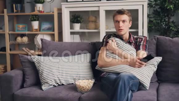 情绪激动的年轻人在家看电视上的恐怖片吃爆米花躲在沙发枕后视频的预览图