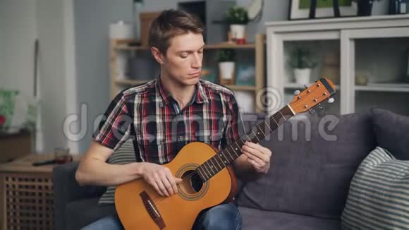 男吉他手正在调音声吉他检查声音触摸琴弦坐在沙发上在家青年文化和视频的预览图