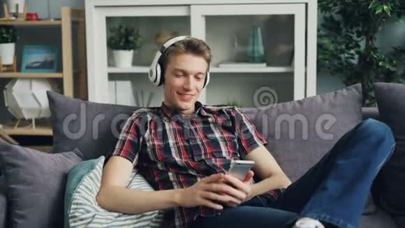 英俊的年轻人正通过耳机听音乐使用智能手机触摸屏幕和大笑视频的预览图