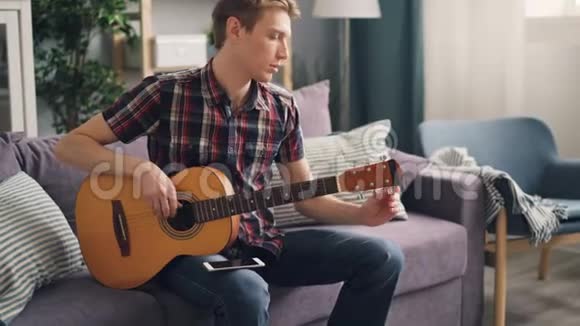 年轻的音乐家正在调整声学吉他在闲暇时间坐在沙发上触摸琴弦青年生活方式视频的预览图