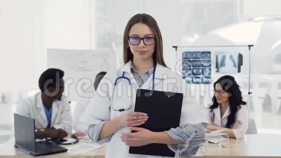 年轻的女医生穿着白色外套听诊器微笑着在诊所的同事背景上看着相机视频的预览图