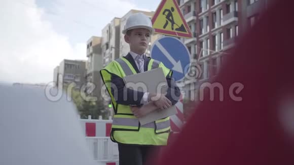 一个有思想的小男孩戴着安全设备和建筑工人头盔站在繁忙的建筑计划上视频的预览图