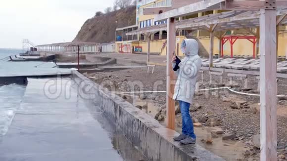 穿白色夹克的女人拿着手机大风浪站在堤岸上侧视视频的预览图