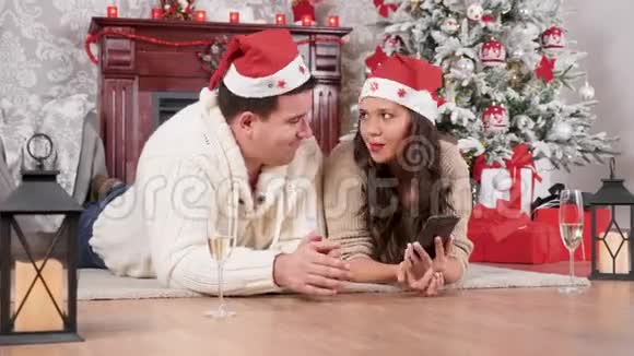 微笑的夫妇坐在地板上用智能手机在线购物圣诞节装饰的房间视频的预览图
