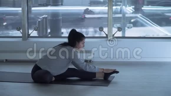 在现代健身中心一个大地板到天花板的健身中心漂亮的黑发女人坐在瑜伽垫上伸展双腿视频的预览图