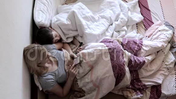 母亲和儿子躺在毯子下面的床上用智能手机搜索互联网写信息玩视频的预览图