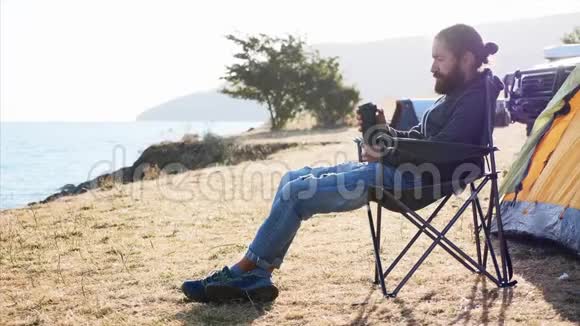 年轻人坐在湖岸露营地的椅子上喝着热水瓶视频的预览图