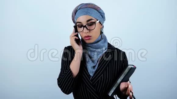 年轻漂亮的阿拉伯商务女性戴着眼镜拿着规划师和笔在智能手机上交谈视频的预览图