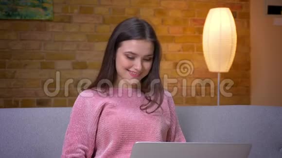 一位年轻漂亮的黑发白种人少女使用笔记本电脑并微笑地坐在沙发上的特写镜头视频的预览图