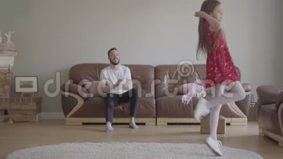 那个穿红裙子的小女孩在蓬松的地毯上的前景上跳舞有胡子的父亲坐在视频的预览图