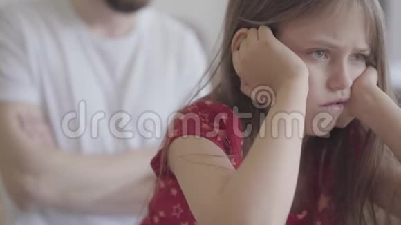 悲伤的小女孩坐在前景上的肖像双手紧紧地抱着她的头父亲交叉双臂视频的预览图