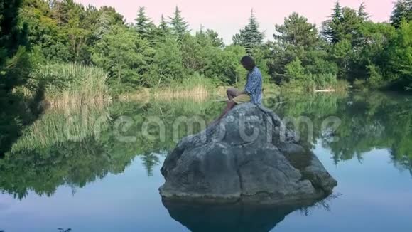 一个年轻人坐在湖中央的石头上自拍使用智能手机进行摄影的概念视频的预览图