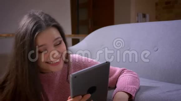在一间舒适的公寓里年轻漂亮的黑发白种人女性在平板电脑上浏览并快乐地笑着视频的预览图