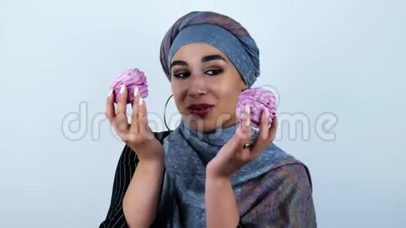 年轻美丽的阿拉伯女人微笑着双手拿着美味的纸杯蛋糕在孤立的白色背景上视频的预览图