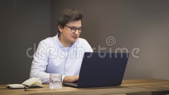 戴眼镜的成年男子坐在桌旁手提电脑上轻敲面带微笑视频的预览图