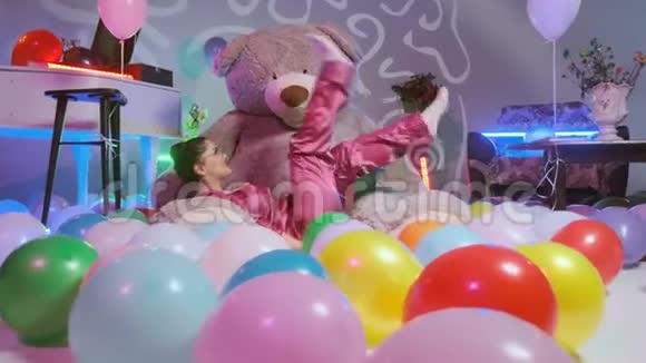 穿着粉红色睡衣的女孩坐在泰迪熊上表现得像一个甜美的女孩看着它微笑着多种颜色视频的预览图