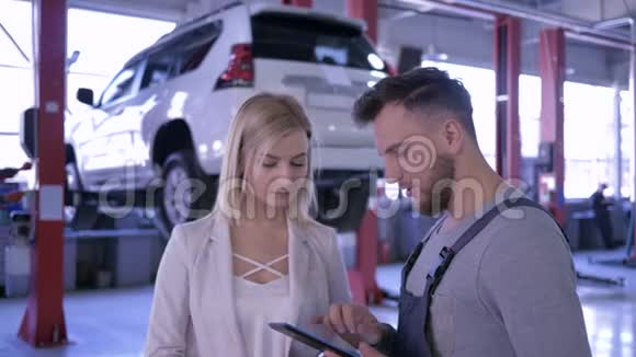汽车修理店带数字平板电脑的机械师在液压升降机上与女客户交谈视频的预览图